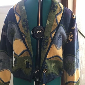 May include: A blue, green, and yellow patterned cardigan with a shawl collar and a decorative button closure. The cardigan has a woven design with stars and snowflakes.