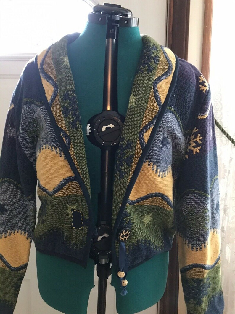 May include: A blue, green, and yellow patterned cardigan with a shawl collar and a decorative button closure. The cardigan has a woven design with stars and snowflakes.