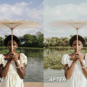 May include: A woman holding a beige paper parasol with a wooden handle. She is wearing a cream-colored dress with puffy sleeves and is standing in front of a body of water with green foliage and trees in the background. The sky is blue with clouds.