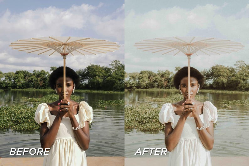May include: A woman holding a beige paper parasol with a wooden handle. She is wearing a cream-colored dress with puffy sleeves and is standing in front of a body of water with green foliage and trees in the background. The sky is blue with clouds.