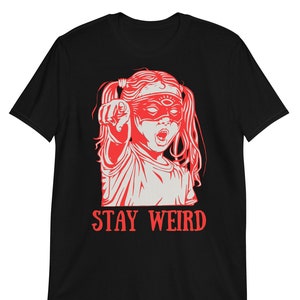 Könnte beinhalten: Schwarzes T-Shirt mit einer roten Illustration eines Mädchens, das eine Maske mit einem Augensymbol trägt. Der Text "STAY WEIRD" ist in Rot unter der Illustration gedruckt.