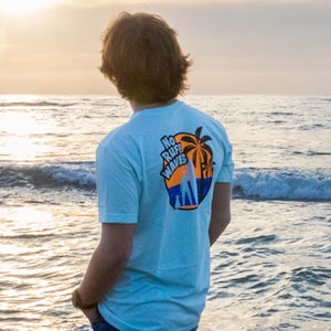 May include: A light blue short-sleeved t-shirt featuring a graphic on the back with a palm tree, a blue ocean wave, and the text "NO RUFF WAVES" in black and orange. The shirt is paired with dark blue denim jeans, creating a casual outfit.