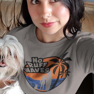 May include: A gray t-shirt with the text "No Ruff Waves" in a circular design featuring a surfer, palm trees, and ocean waves. The shirt is paired with blue jeans.