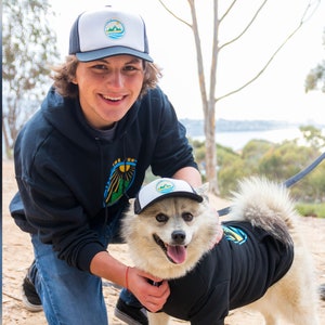 A man and his dog in nature. They are wearing matching dog and owner trucker hats from PupLid and Fetch the Sun. The hats have a logo printed on the white foam.