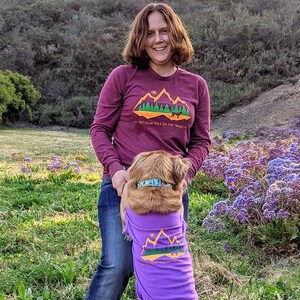 Woman wearing a maroon long sleeve shirt with her dog in a purple dog shirt for dogs. Both have matching hiking graphics