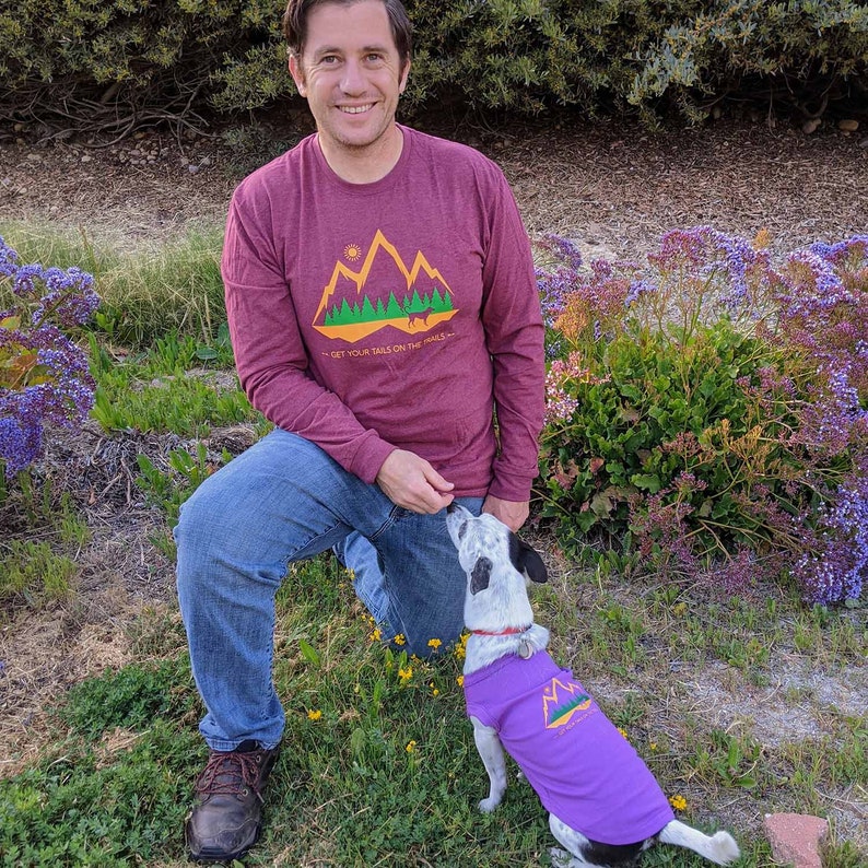man and his dog in a field wearing matching dog and owner shirts. His shirt is maroon long sleeve and the dog is wearing purple.