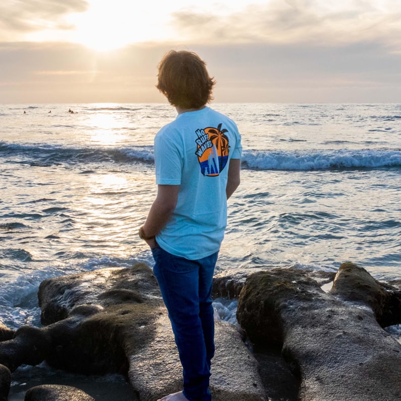 May include: A light blue short-sleeved t-shirt featuring a graphic on the back with a palm tree, a blue ocean wave, and the text "NO RUFF WAVES" in black and orange. The shirt is paired with dark blue denim jeans, creating a casual outfit.