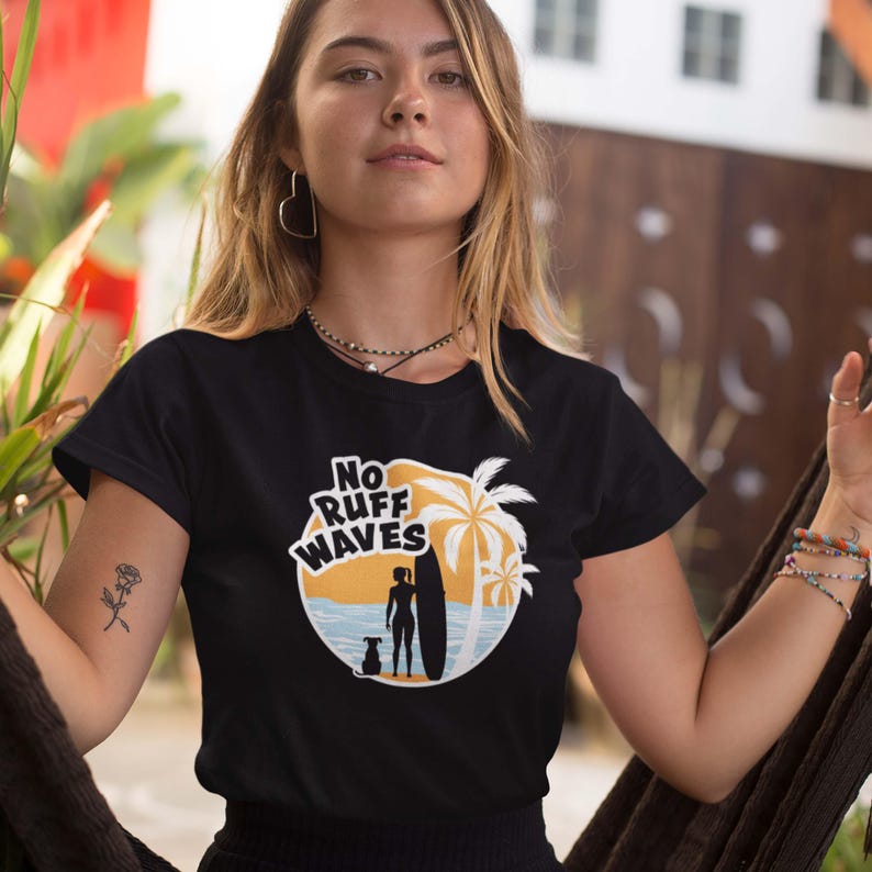 May include: A black short-sleeved t-shirt featuring a circular graphic with a beach scene, a palm tree, a person holding a surfboard, and a dog. The text "NO RUFF WAVES" is printed in bold white letters above the silhouette of the person and dog.
