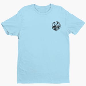May include: A light blue short-sleeve t-shirt featuring a circular black graphic on the upper left chest. The graphic depicts a mountain landscape with a sun and a dog, surrounded by the text "FETCH THE SUN" in a circular border.