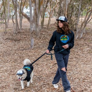 A woman hiking with her dog. They are wearing matching dog and owner trucker hats from Fetch the Sun and PupLid. The hats are black. They are also wearing matching dog and owner hoodies.