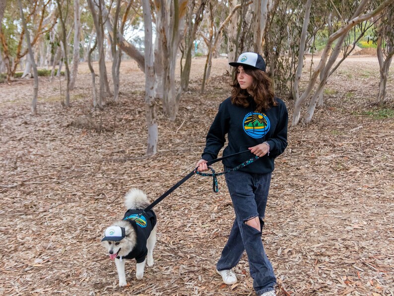 A woman hiking with her dog. They are wearing matching dog and owner trucker hats from Fetch the Sun and PupLid. The hats are black. They are also wearing matching dog and owner hoodies.