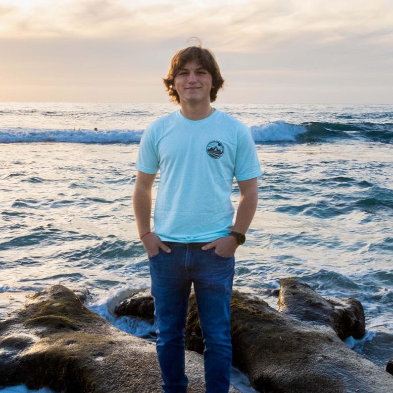 May include: A light blue short-sleeve t-shirt featuring a small circular graphic on the left chest, paired with dark blue denim jeans. The t-shirt has a crew neckline and a simple, solid-colored design, worn with a dark-colored wristwatch on the wrist.