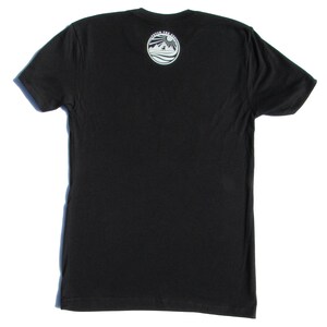 May include: A black short-sleeved t-shirt featuring a circular white graphic on the upper back. The graphic depicts a sun, mountains, and a dog, with the text "FETCH THE SUN" arched along the top edge of the circle. The shirt has a crew neckline.