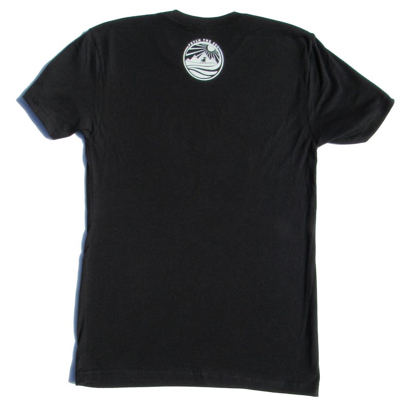 May include: A black short-sleeved t-shirt featuring a circular white graphic on the upper back. The graphic depicts a sun, mountains, and a dog, with the text "FETCH THE SUN" arched along the top edge of the circle. The shirt has a crew neckline.