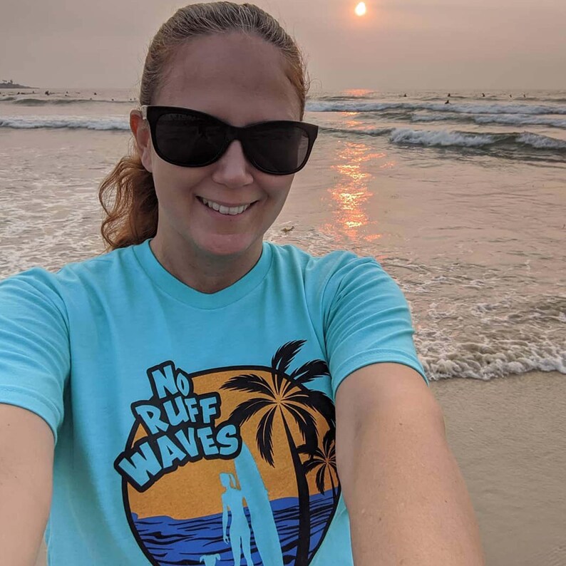 May include: A light blue t-shirt featuring a graphic print of a sunset, palm trees, a surfboard, and a person with a dog. The text "NO RUFF WAVES" is printed in bold blue letters over the orange sunset circle design on the front of the shirt.