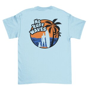May include: A light blue t-shirt featuring a circular graphic on the back. The design shows a silhouette of a person with a surfboard and a dog standing on a beach with palm trees, an orange sun, and blue ocean waves, with the text No Ruff Waves.