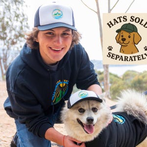 Puede incluir: Gorra de camionero blanca y azul marino con un logotipo de montaña y olas. La gorra la lleva una persona y un perro, ambos con ropa a juego. La imagen incluye un letrero que dice "Sombreros se venden por separado".