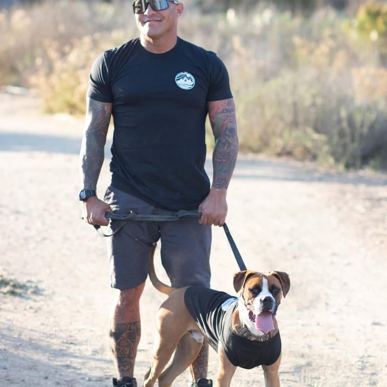 May include: A black short-sleeved t-shirt with a circular logo on the left chest featuring a mountain graphic and text. The shirt is worn with dark gray shorts. A brown and white dog stands in front wearing a matching black sleeveless shirt.