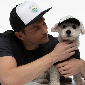 A man and his small dog in a photo studio. They are wearing matching dog and owner trucker hats. The hats are black. They have a Fetch the Sun logo printed on them in blue, green, and yellow.