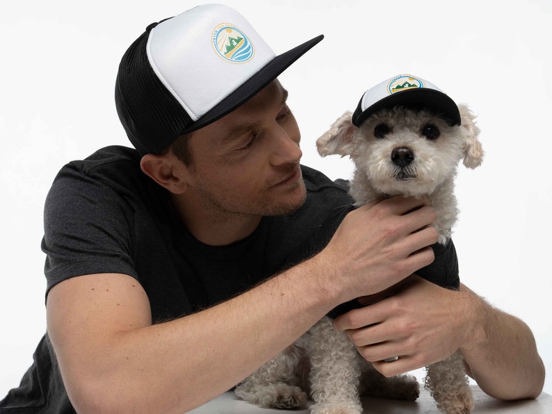 A man and his small dog in a photo studio. They are wearing matching dog and owner trucker hats. The hats are black. They have a Fetch the Sun logo printed on them in blue, green, and yellow.