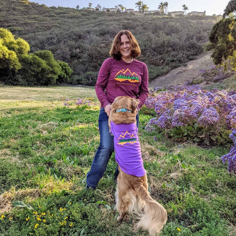 Woman wearing a maroon long sleeve shirt with her dog in a purple dog shirt for dogs. Both have matching hiking graphics