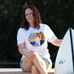 May include: A white short-sleeved t-shirt featuring a circular graphic on the chest. The design depicts a sunset over the ocean with palm trees, a silhouette of a person, and a dog, accompanied by the text No Ruff Waves in bold blue letters.