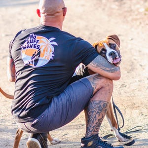 May include: A black short-sleeved t-shirt featuring a circular graphic on the back with the text NO RUFF WAVES, a silhouette of a person surfing, and palm trees against an orange sun. The shirt is paired with dark gray shorts and black sneakers.