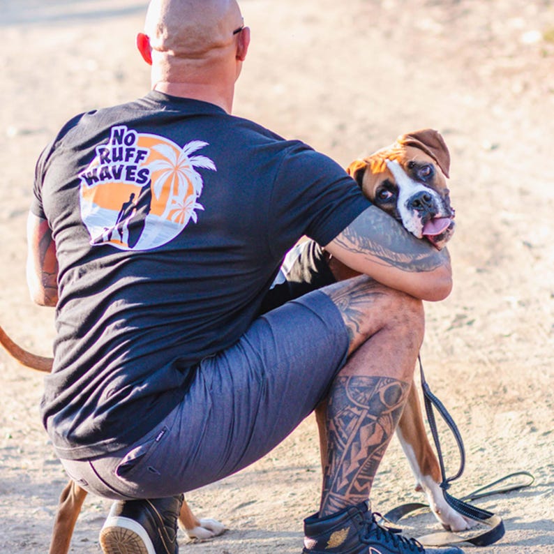 May include: A black short-sleeved t-shirt featuring a circular graphic on the back with the text NO RUFF WAVES, a silhouette of a person surfing, and palm trees against an orange sun. The shirt is paired with dark gray shorts and black sneakers.