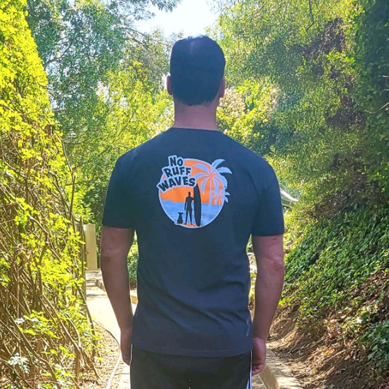 May include: A dark navy blue short-sleeve t-shirt featuring a circular graphic on the back. The design depicts a silhouette of a person with a surfboard and a dog standing on a beach, with palm trees and the text "NO RUFF WAVES" in white.