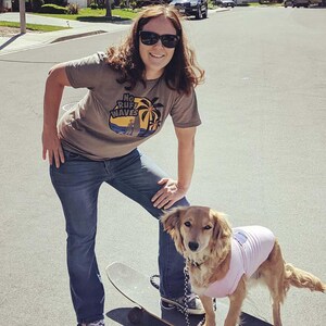 May include: A taupe short-sleeved t-shirt featuring a circular graphic with palm trees, a surfboard, and the text "NO RUFF WAVES" in black and yellow. The shirt is paired with blue denim jeans and worn with black sunglasses.