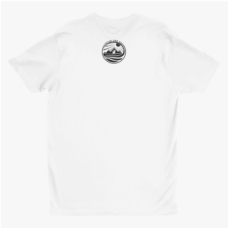May include: A plain white short-sleeved t-shirt featuring a circular black graphic on the upper back. The design depicts a dog silhouette standing before mountains and a sun, with the text FETCH THE SUN arched along the top of the circle.