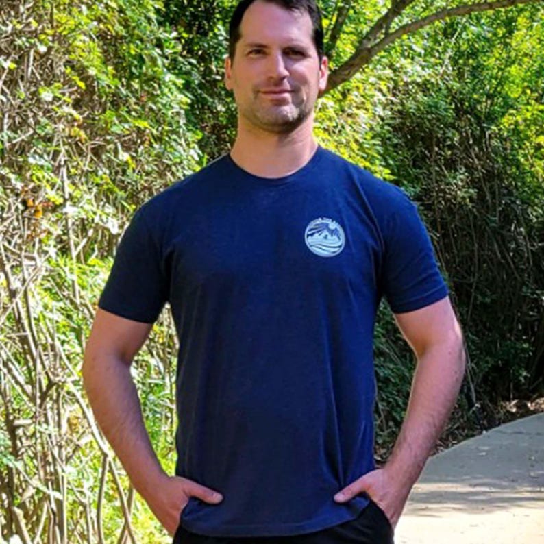 May include: A dark blue short-sleeved t-shirt featuring a circular graphic on the left chest. The graphic depicts a stylized mountain range and sun design with the text "PEAK AND PINE" arched along the top edge of the circular emblem.