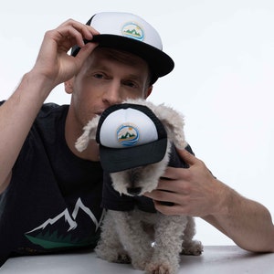 A man and his dog wearing matching dog and owner trucker hats. The hats are black and have a Fetch the Sun adventure dog printed on the front. The front is white foam and the mesh and bill are black.