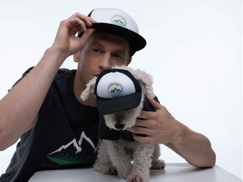 A man and his dog wearing matching dog and owner trucker hats. The hats are black and have a Fetch the Sun adventure dog printed on the front. The front is white foam and the mesh and bill are black.