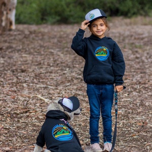 A dog loving kid and her dog wearing matching dog and owner hats and hoodies. The hat has a white foam front and has a black bill and black mesh. There is a Fetch the Sun logo printed on the front and the logo colors are blue, green, and yellow.