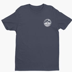 May include: A dark navy blue short-sleeve t-shirt featuring a circular white graphic on the upper left chest. The graphic depicts a dog standing before mountains and a sun, with the text "FETCH THE SUN" arched along the top edge of the circle.