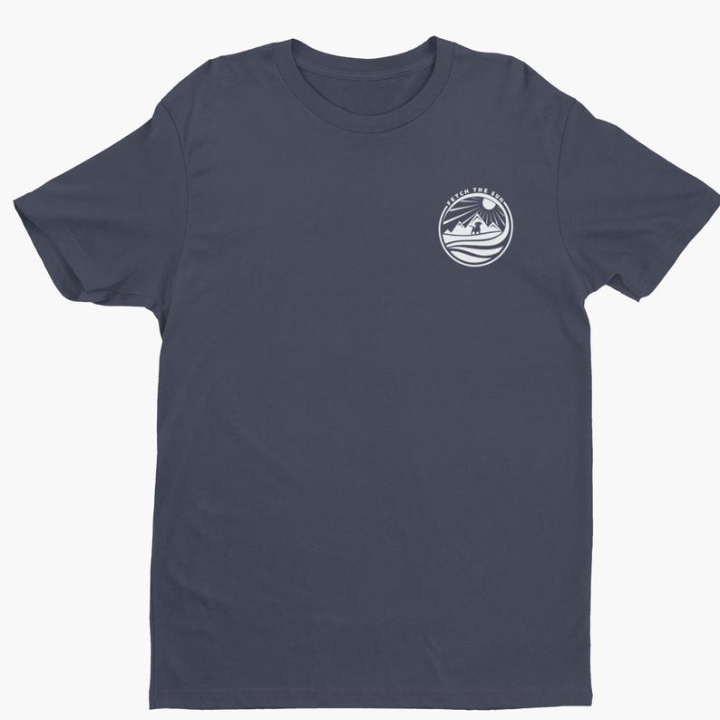 May include: A dark navy blue short-sleeve t-shirt featuring a circular white graphic on the upper left chest. The graphic depicts a dog standing before mountains and a sun, with the text "FETCH THE SUN" arched along the top edge of the circle.