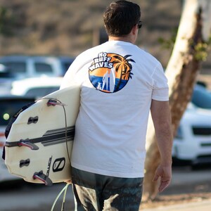 May include: A white short-sleeve t-shirt featuring a circular graphic on the back with the text NO RUFF WAVES, depicting a silhouette of a person with a surfboard and a dog against a sunset, orange sky, and blue ocean with palm trees.