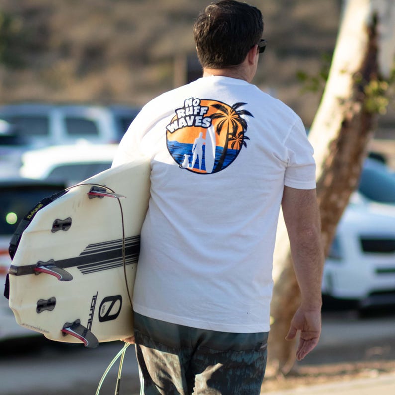 May include: A white short-sleeve t-shirt featuring a circular graphic on the back with the text NO RUFF WAVES, depicting a silhouette of a person with a surfboard and a dog against a sunset, orange sky, and blue ocean with palm trees.