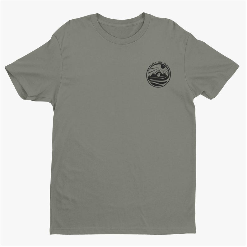 May include: A sage green short-sleeve t-shirt featuring a circular black graphic on the left chest. The graphic depicts a mountain landscape with a sun and waves, surrounded by the text CATCH THE SUN in a circular border.