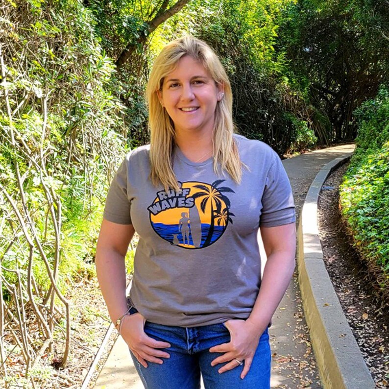 May include: A grey short-sleeved t-shirt featuring a circular graphic on the chest. The graphic depicts a yellow sunset, blue ocean waves, black palm trees, and a silhouette of a person with a surfboard and a dog, with the text RUFF WAVES.