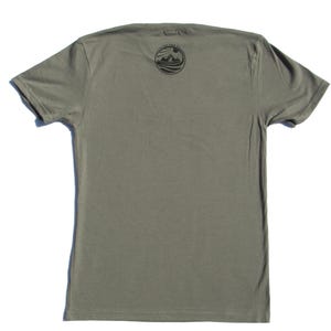 May include: A short-sleeved olive green t-shirt featuring a circular black graphic on the upper chest area. The graphic depicts a sun rising over mountains and water with the text "CATCH THE SUN" arched along the top edge of the circle.