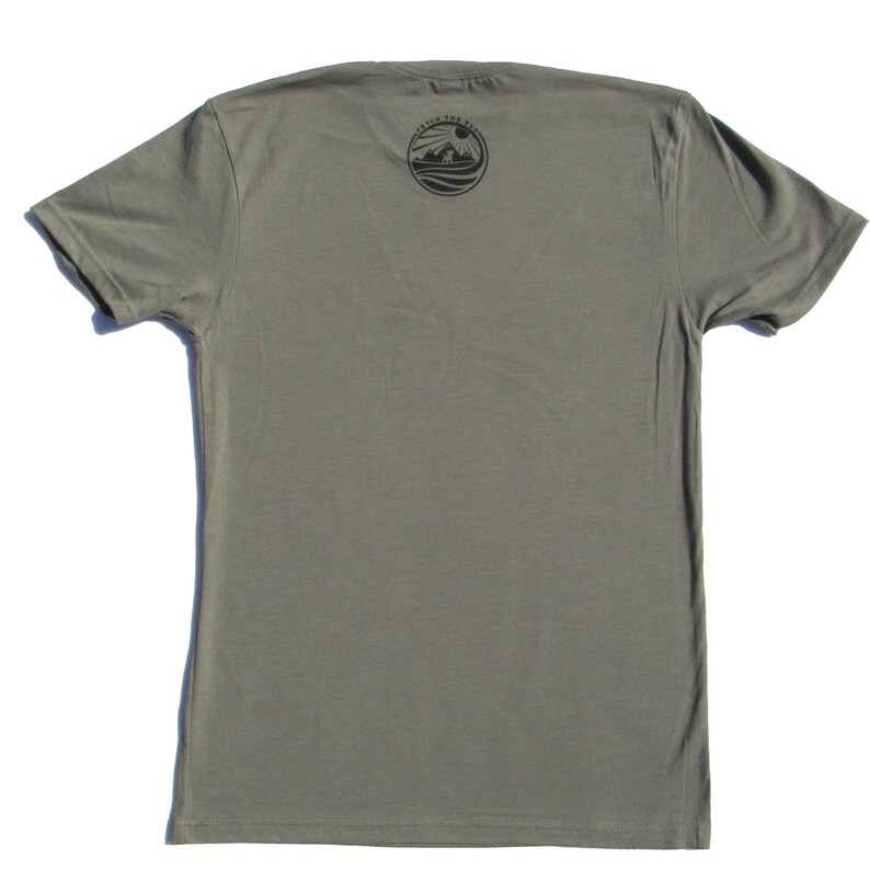 May include: A short-sleeved olive green t-shirt featuring a circular black graphic on the upper chest area. The graphic depicts a sun rising over mountains and water with the text "CATCH THE SUN" arched along the top edge of the circle.