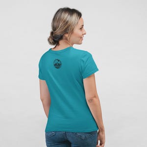 May include: A short-sleeved teal t-shirt featuring a small, circular black graphic on the upper back. The graphic depicts a landscape scene with a sun, mountains, and water, enclosed within a circular border with text along the edge.