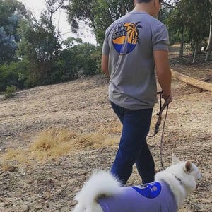 May include: A gray short-sleeved t-shirt featuring a circular graphic on the back with palm trees, ocean waves, and the text NO RUFF WAVES. The shirt is paired with dark blue jeans and a light purple dog shirt with a matching circular graphic.