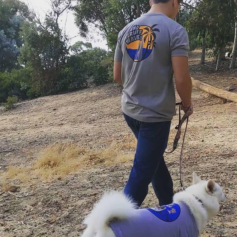 May include: A gray short-sleeved t-shirt featuring a circular graphic on the back with palm trees, ocean waves, and the text NO RUFF WAVES. The shirt is paired with dark blue jeans and a light purple dog shirt with a matching circular graphic.
