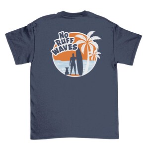 May include: A navy blue short-sleeve t-shirt featuring a circular graphic on the back. The design shows a silhouette of a person with a surfboard and a dog standing on a beach with palm trees against an orange sun. The text reads NO RUFF WAVES.