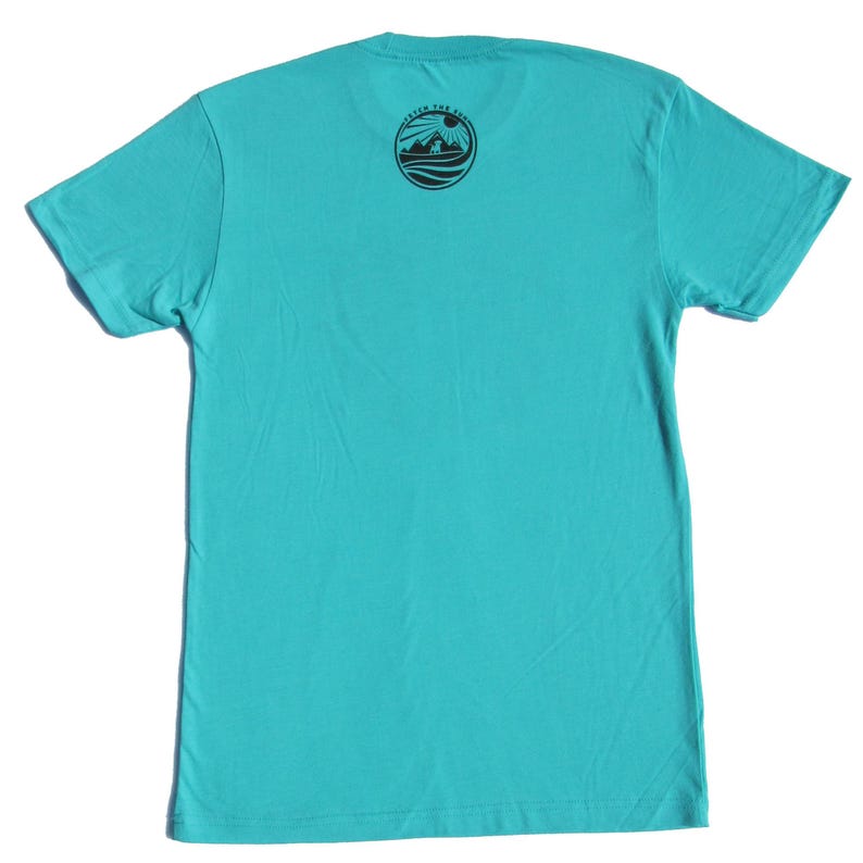 May include: A bright turquoise short-sleeve t-shirt featuring a circular black graphic on the upper back. The graphic depicts a mountain landscape with a sun and the text CATCH THE SUN in a circular border. The shirt has a crew neckline.