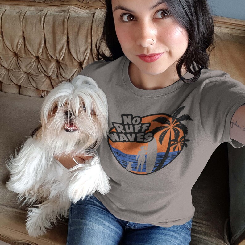 May include: A gray t-shirt with the text "No Ruff Waves" in a circular design featuring a surfer, palm trees, and ocean waves. The shirt is paired with blue jeans.