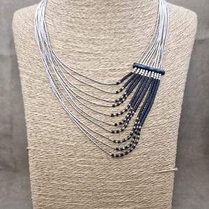 May include: A silver-toned necklace with multiple strands cascading down. The necklace features a dark blue rectangular accent with black beads and silver beads, creating a layered effect. The jewelry is displayed on a mannequin.